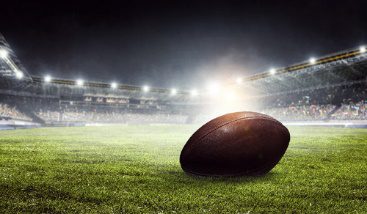 A football on the grass in front of a stadium.